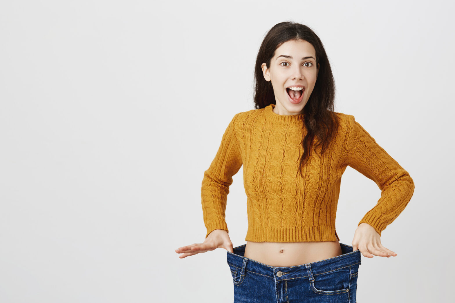 Astonished and happy young lady being impressed and excited because of losing weight, showing empty space in jeans by stretching it, standing over gray background. Girl is ready for summer